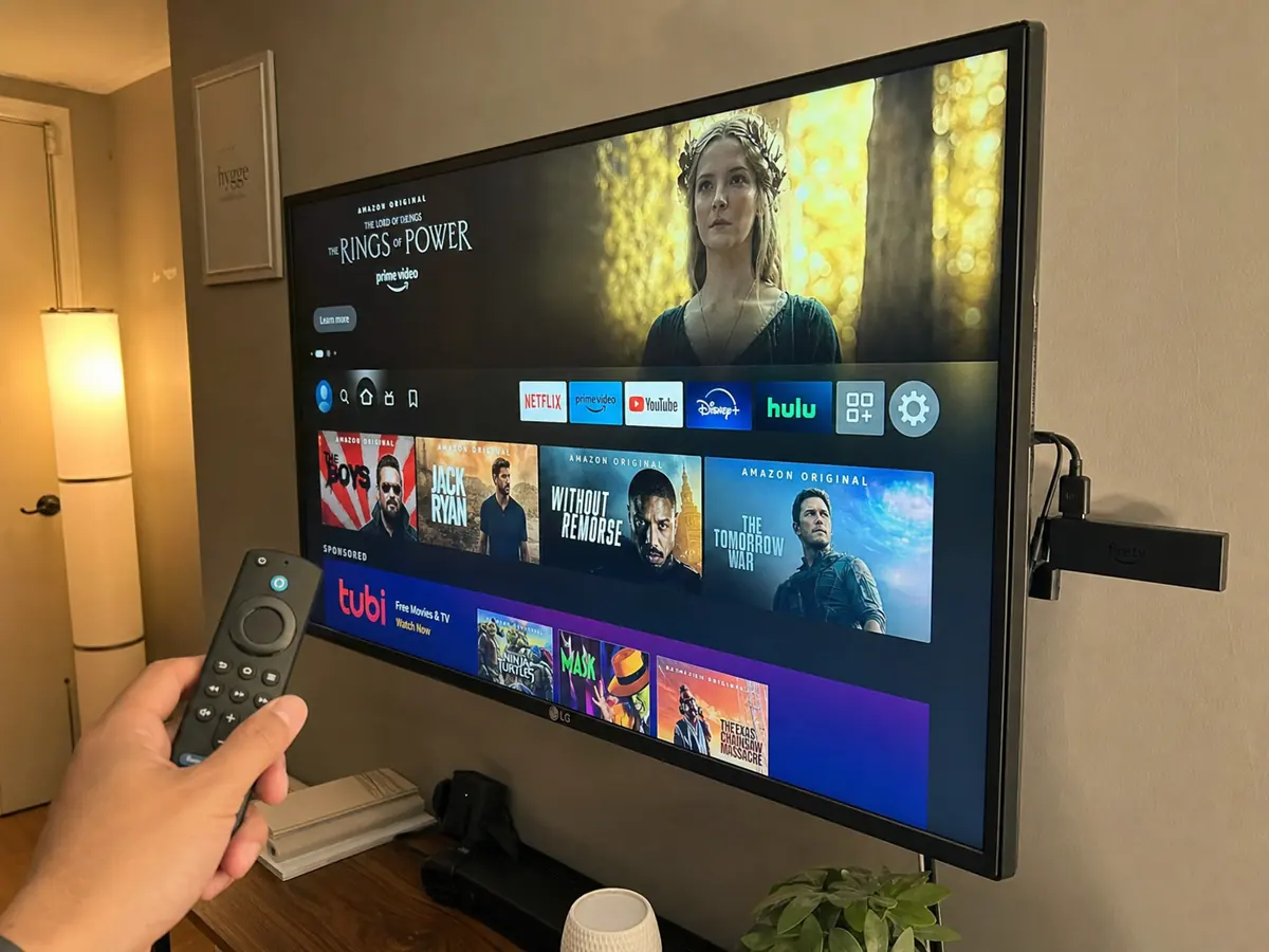 Fire Stick conectado atrás de Smart TV LG com mão segurando controle remoto Alexa